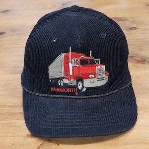 May include: A navy blue corduroy baseball cap featuring an embroidered graphic of a red semi-truck with a gray trailer. The word KENWORTH is stitched in red capital letters beneath the truck. The cap has a structured crown and a curved brim.