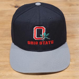 Peut inclure: Une casquette de baseball bleu marine avec une visière plate grise et un bouton supérieur gris. L'avant présente une lettre O rouge brodée avec une feuille de marronnier verte et le texte OHIO STATE brodé en rouge en dessous.