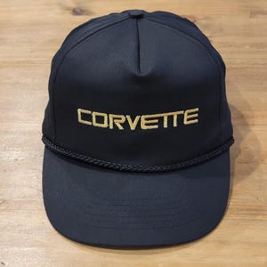 May include: A black baseball cap featuring the word CORVETTE embroidered in gold thread across the front panel. The cap includes a matching black braided cord trim positioned horizontally across the base of the crown, just above the flat brim.