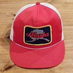 May include: A red and white trucker hat featuring a rectangular patch on the front. The patch has a dark blue background with a silver map of Alaska and the word Alaska embroidered in red script. The hat has a red brim and white mesh back.