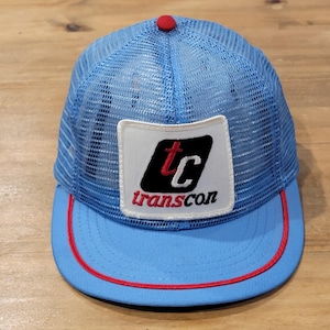 May include: A light blue trucker hat featuring a mesh back and a solid front panel. A white rectangular patch on the front displays a black logo with the red letters tc and the word transcon in red. The hat has a red button on top and red trim.