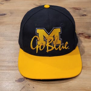 May include: A navy blue baseball cap with a yellow brim and a yellow button on top. The front features an embroidered yellow block letter M with the yellow cursive text Go Blue underneath. The design includes small ventilation eyelets on the crown.