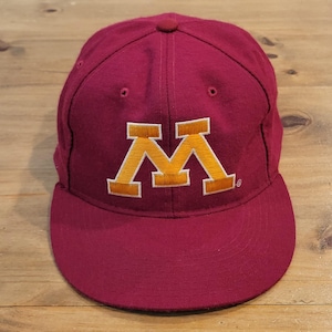 May include: A maroon baseball cap featuring a prominent embroidered block letter M in gold with a white outline on the front panel. The cap has a structured crown, a flat brim, and small ventilation eyelets stitched into the top fabric panels.