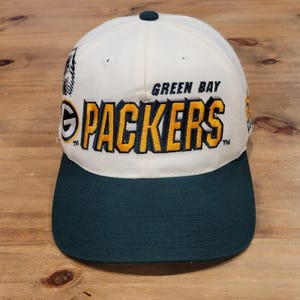 以下が含まれることがあります： クリーム色のベースボールキャップ。前面には黄色い刺繍で「GREEN BAY PACKERS」の文字がダークグリーンの縁取りで施されています。ダークグリーンのつば、頂部のダークグリーンのボタン、前面左側に小さなロゴグラフィックが特徴です。