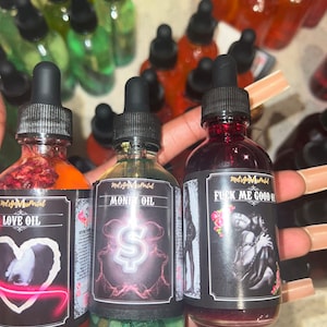 May include: Three glass bottles of oil with black droppers. The bottles have labels with the text "LOVE OIL", "MONEY OIL", and "FUCK ME GOOD OIL". The "MONEY OIL" bottle has green glitter at the bottom. The oils are red, orange, and green.