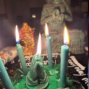 May include: A spiritual arrangement featuring lit teal candles, including one shaped like praying hands. A golden Buddha statue and a green statue are in the background. The candles are on a black surface with white script and green powder.