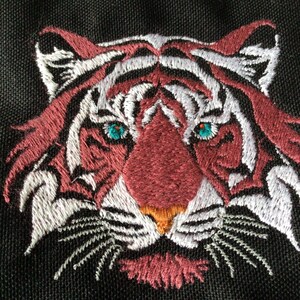 May include: Embroidered tiger face in red, white, and black thread on a black background. The tiger has teal eyes and an orange nose. The detailed embroidery creates a textured and striking design.
