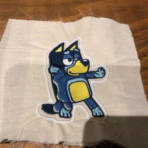 May include: Embroidered patch of a cartoon dog in blue, yellow, and black. The patch is on a square of off-white fabric with frayed edges. The dog is in a dynamic pose, with one arm raised. The patch is suitable for clothing.