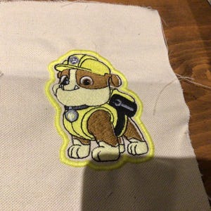 May include: Embroidered patch of a cartoon dog wearing a yellow hard hat and vest. The dog is brown and white with a black backpack and a yellow outline. The patch is on a beige fabric.