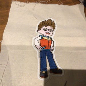 May include: Embroidered patch of a cartoon character in a red and orange vest, blue trousers, and black shoes. The patch is on a piece of beige fabric, with visible stitching around the edges. The character has brown hair and is smiling.