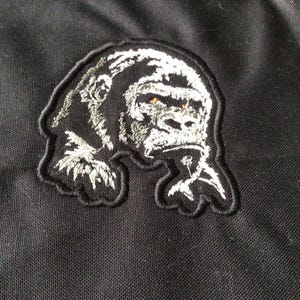 May include: Embroidered patch with a gorilla design. The patch has a black border and a detailed white and grey gorilla image with orange eyes. It is set against a black fabric background.