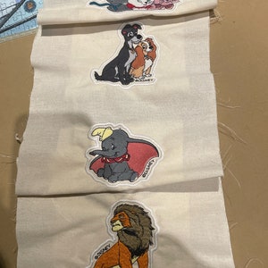 May include: Embroidered patches featuring Disney characters: Marie and her siblings, Lady and Tramp, Dumbo, and Scar. Each patch is on a cream-coloured fabric, with the Disney logo visible. Suitable for crafting or customising clothing.