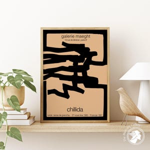 May include: A framed poster with a black and white abstract design on a peach background. The poster has the text 'galerie maeght', '13 rue de téhéran, paris 8', 'chillida', 'lumak-terres de grand feu 27 novembre 1980-15 jenvier 1981'.