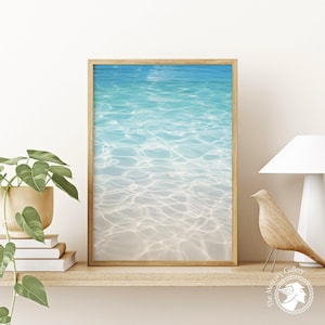 Può includere: Una stampa incorniciata raffigurante una serena scena di spiaggia con acqua turchese e limpida. L'opera cattura il gioco di luci e ombre sulla superficie dell'acqua, creando un senso di profondità e tranquillità. La cornice è di colore legno chiaro.