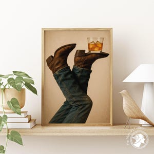 Puede incluir: Una impresión enmarcada que presenta un par de piernas con vaqueros azules y botas de vaquero marrones, sosteniendo un vaso de whisky con hielo. La obra de arte se exhibe en una estantería de madera con una planta, libros, una lámpara y una figura de pájaro de madera.