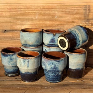 Peut inclure: Ensemble de tasses en céramique faites à la main avec une esthétique rustique. Les tasses présentent un glaçage bleu clair avec des accents bleu foncé et marron. L'intérieur des tasses est de couleur marron chaud. Les tasses sont empilées sur une surface en bois.
