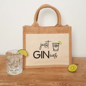 Könnte beinhalten: Eine braune Jute-Tragetasche mit einem weißen Canvas-Einsatz. Der Canvas-Einsatz hat schwarzen Text, der "just GINius" lautet, und eine schwarze Strichzeichnung eines Gin-Glases mit einem Limettenkeil.