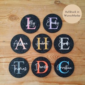Könnte beinhalten: Neun dunkelgraue Filzuntersetzer mit personalisierten Namen und Initialen in verschiedenen Farben. Die Namen sind in weißer Schrift, und die Initialen sind in Rosa, Lila, Pfirsich, Gold, Grau, Orange und Blau. Der Text "Aufdruck in Wunschfarbe" befindet sich in einem hellbraunen Kreis.