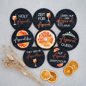 May include: Set of eight round black felt coasters with various designs and text related to Aperol, an Italian liqueur. The designs include an orange slice, a glass of Aperol spritz, and text such as "Holy Aperol", "Zeit f&uuml;r Aperol", "Aperol Queen", and "Aperol Spritz Tour".