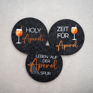 May include: Three black felt coasters with white text and orange graphics. The coasters feature the text "Holy Aperol", "Zeit f&uuml;r Aperol", and "Leben auf der Aperol Spur".