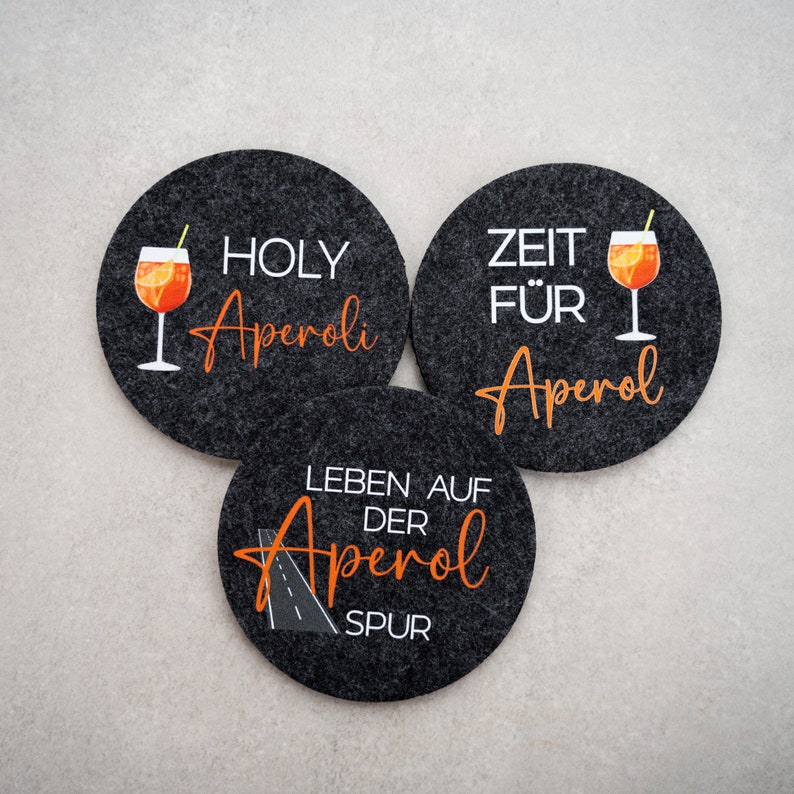 May include: Three black felt coasters with white text and orange graphics. The coasters feature the text "Holy Aperol", "Zeit f&uuml;r Aperol", and "Leben auf der Aperol Spur".