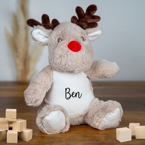May include: A plush reindeer toy with a red nose and brown antlers. The reindeer is wearing a white shirt with the name "Ben" embroidered on it.
