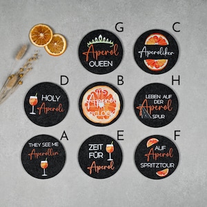 May include: Ten black round coasters with various designs and text related to Aperol, an Italian liqueur. The designs include a glass of Aperol spritz, an orange slice, and text such as "Holy Aperoli", "It's Aperol O'Clock", and "Zeit f&uuml;r Aperol".