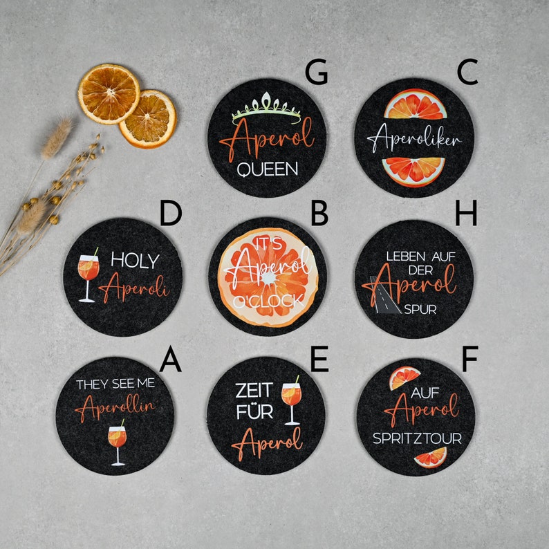 May include: Ten black round coasters with various designs and text related to Aperol, an Italian liqueur. The designs include a glass of Aperol spritz, an orange slice, and text such as "Holy Aperoli", "It's Aperol O'Clock", and "Zeit f&uuml;r Aperol".