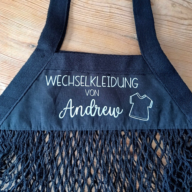 May include: Black mesh bag with a black fabric top. The fabric top has white text that says "Wechselkleidung von Andrew" and a white outline of a t-shirt.