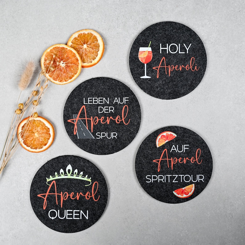 May include: Four black round coasters with different designs featuring the text "Aperol" in white and orange. The coasters have different designs including a glass of Aperol Spritz, a road with the text "Leben auf der Aperol Spur", a crown with the text "Aperol Queen", and an orange slice with the text "Auf Aperol Spritztour".