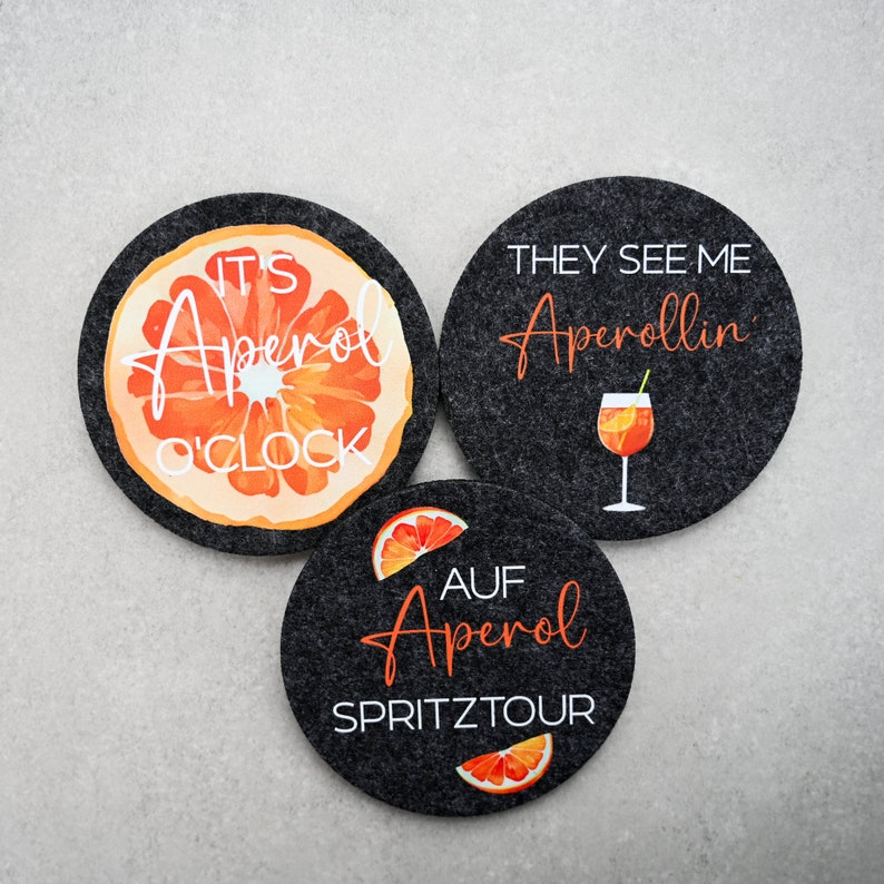 May include: Three black felt coasters with orange and white designs. The first coaster says "It's Aperol O'Clock" and features a slice of orange. The second coaster says "They See Me Aperollin'" and features a glass of Aperol Spritz. The third coaster says "Auf Aperol Spritztour" and features a slice of orange.