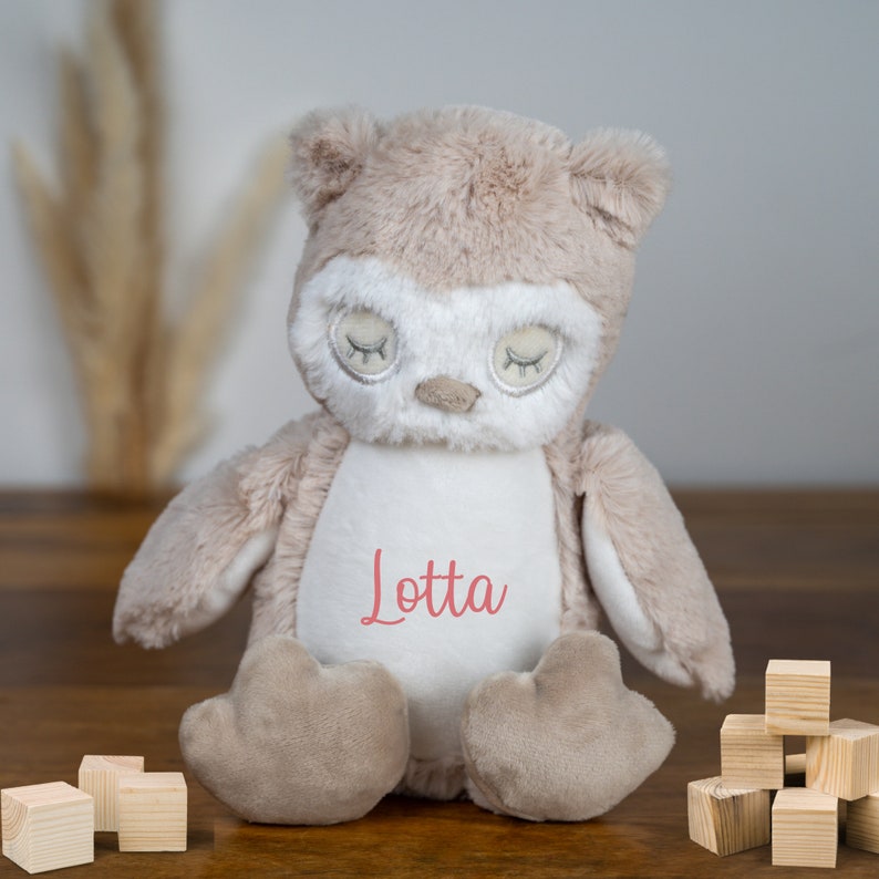 May include: A soft, plush owl toy with a white belly and closed eyes. The owl is sitting on a wooden surface and has the name "Lotta" embroidered on its chest.