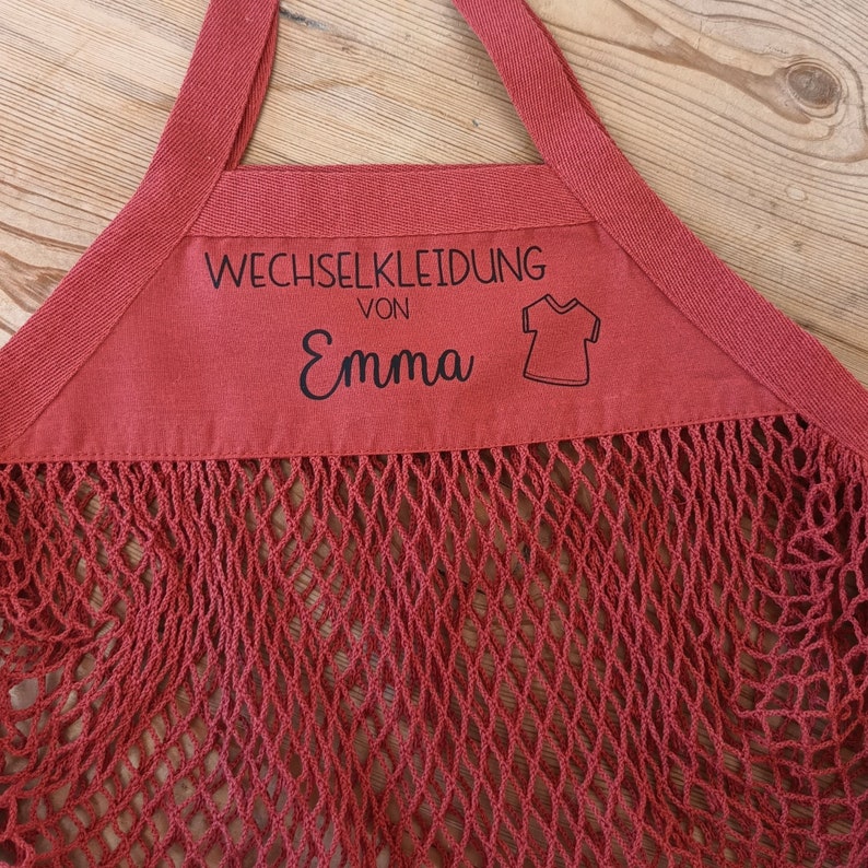 May include: Red mesh bag with a red fabric top. The fabric top has a black label that says "Wechselkleidung von Emma" with a small black illustration of a t-shirt.