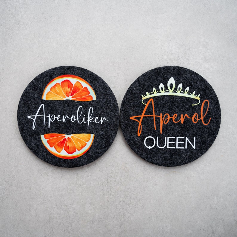 May include: Two black felt coasters with white text. One coaster features an orange slice and the text "Aperoliker". The other coaster features a crown and the text "Aperol Queen".