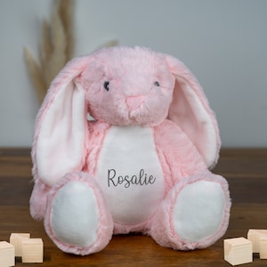 May include: Pink plush bunny rabbit with white ears and a white patch on its chest. The bunny has black eyes and a stitched nose. The bunny is sitting on a wooden surface and has the name "Rosalie" embroidered on its chest.