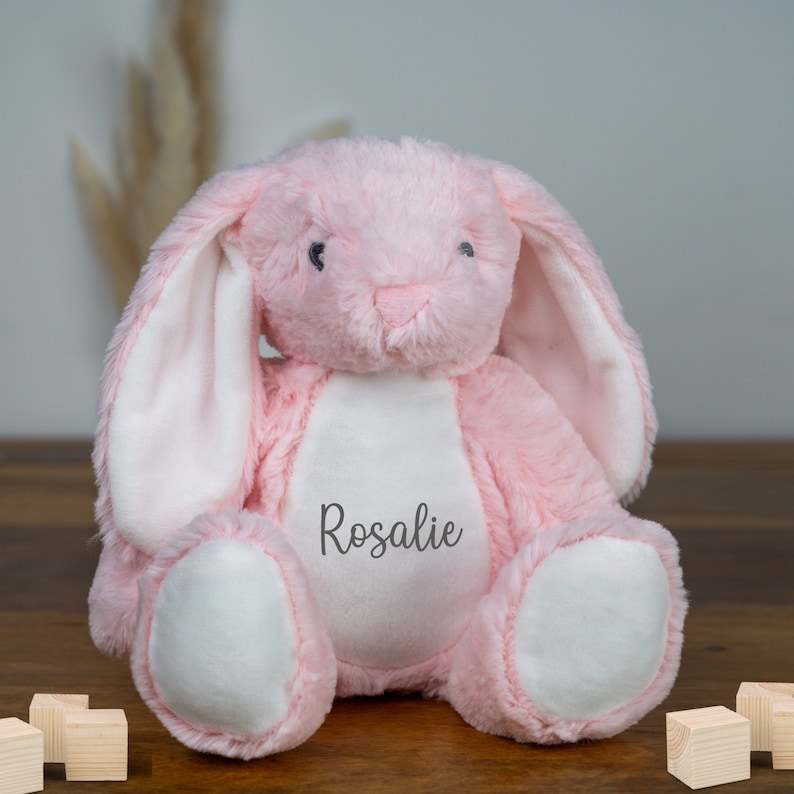 May include: Pink plush bunny rabbit with white ears and a white patch on its chest. The bunny has black eyes and a stitched nose. The bunny is sitting on a wooden surface and has the name "Rosalie" embroidered on its chest.