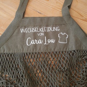 May include: A green canvas bag with a mesh bottom. The bag has a white label that says "Wechselkleidung von Cara Lou" and a small graphic of a t-shirt.