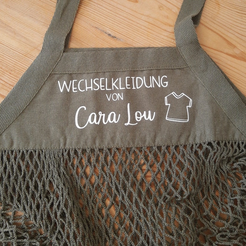 May include: A green canvas bag with a mesh bottom. The bag has a white label that says "Wechselkleidung von Cara Lou" and a small graphic of a t-shirt.