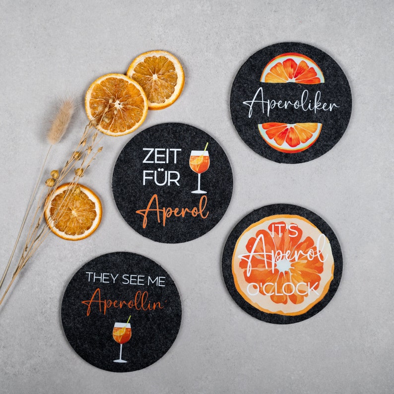 May include: Four black coasters with orange and white designs featuring the text "Aperol" and "Aperoliker". One coaster says "Zeit f&uuml;r Aperol", another says "They see me Aperollin", and the last one says "It's Aperol o'clock".