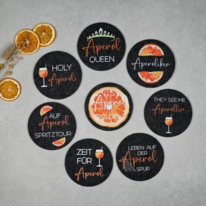 May include: Eight black round coasters with orange and white designs featuring the text "Aperol" and various phrases related to the Italian aperitivo drink. The coasters are made of felt and have a smooth surface.