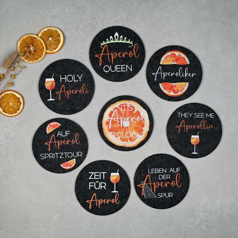 May include: Eight black round coasters with orange and white designs featuring the text "Aperol" and various phrases related to the Italian aperitivo drink. The coasters are made of felt and have a smooth surface.