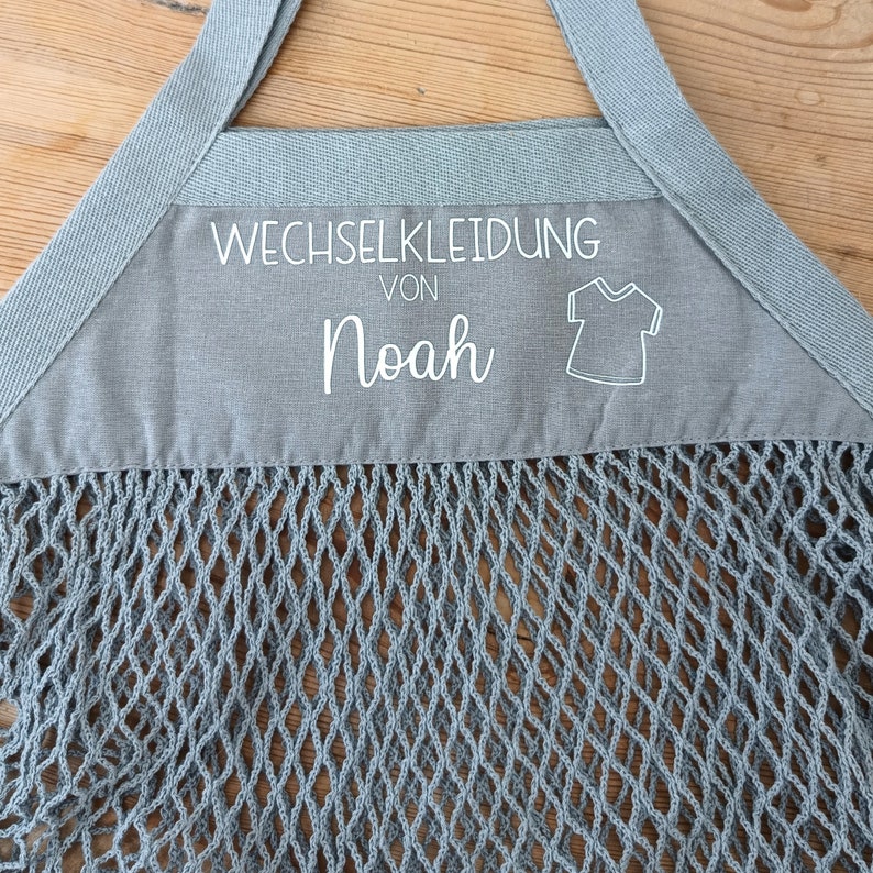 May include: A gray mesh bag with a gray fabric top. The fabric top has white text that says "Wechselkleidung von Noah" and a white outline of a t-shirt.