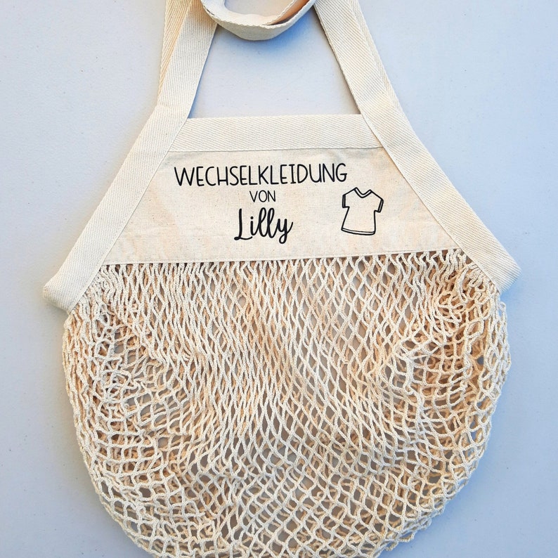 May include: A beige canvas tote bag with a mesh bottom. The bag has a white label that says "Wechselkleidung von Lilly" and a small graphic of a t-shirt.