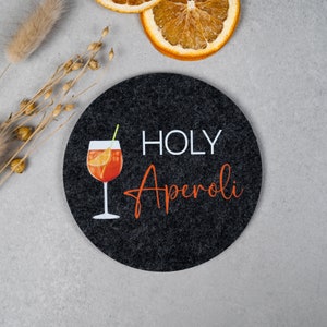 May include: Black round coaster with the text "HOLY Aperol" in white and orange lettering. A glass of Aperol Spritz is pictured on the coaster.