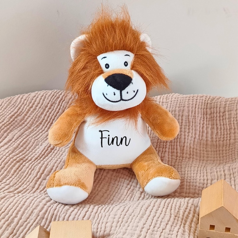 May include: A plush lion toy with a brown and white fur, a black nose, and a smiling face. The toy has a white patch on its chest with the name "Finn" embroidered in black.