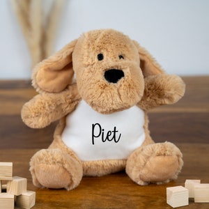 May include: A brown plush dog toy with a white shirt that says "Piet" on it. The toy is sitting on a wooden surface.