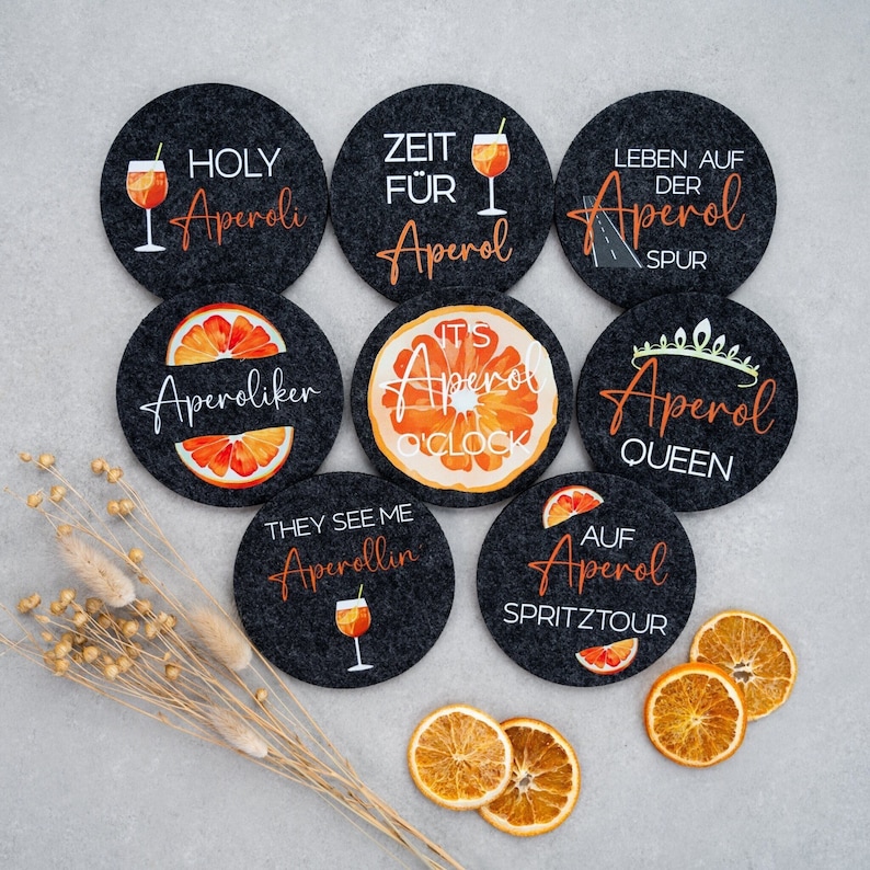 May include: Set of eight round black felt coasters with various designs and text related to Aperol, an Italian liqueur. The designs include an orange slice, a glass of Aperol spritz, and text such as "Holy Aperol", "Zeit f&uuml;r Aperol", "Aperol Queen", and "Aperol Spritz Tour".