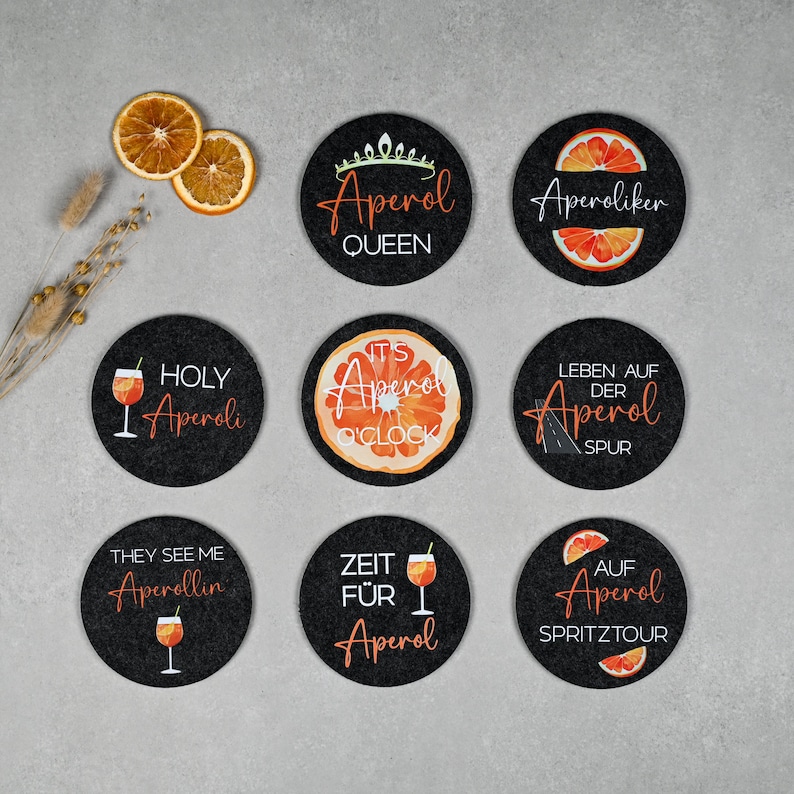 May include: Set of eight black round coasters with orange and white designs featuring the word "Aperol" and various phrases. The coasters have a variety of designs, including a glass of Aperol spritz, an orange slice, and a road sign.