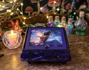 May include: A square wooden box with a dark purple finish featuring a starry night pattern. The lid displays a printed image of a fairy with iridescent wings sitting in a landscape under a crescent moon. The box has a small black knob and feet.
