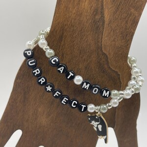 May include: Two beaded bracelets with black letter beads spelling "Cat Mom" and "Purrfect" with a small black and white cat charm hanging from the "Purrfect" bracelet.